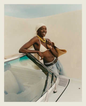 Smiling Black woman in bikini and headscarf leaning against car on sand dune. Car is cream-colored, with an older vintage aesthetic.