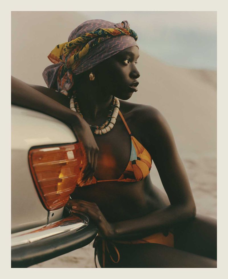 Adult woman in orange bikini and patterned headscarf leaning on a vintage car in an outdoor, sunlit desert setting with sand dunes.