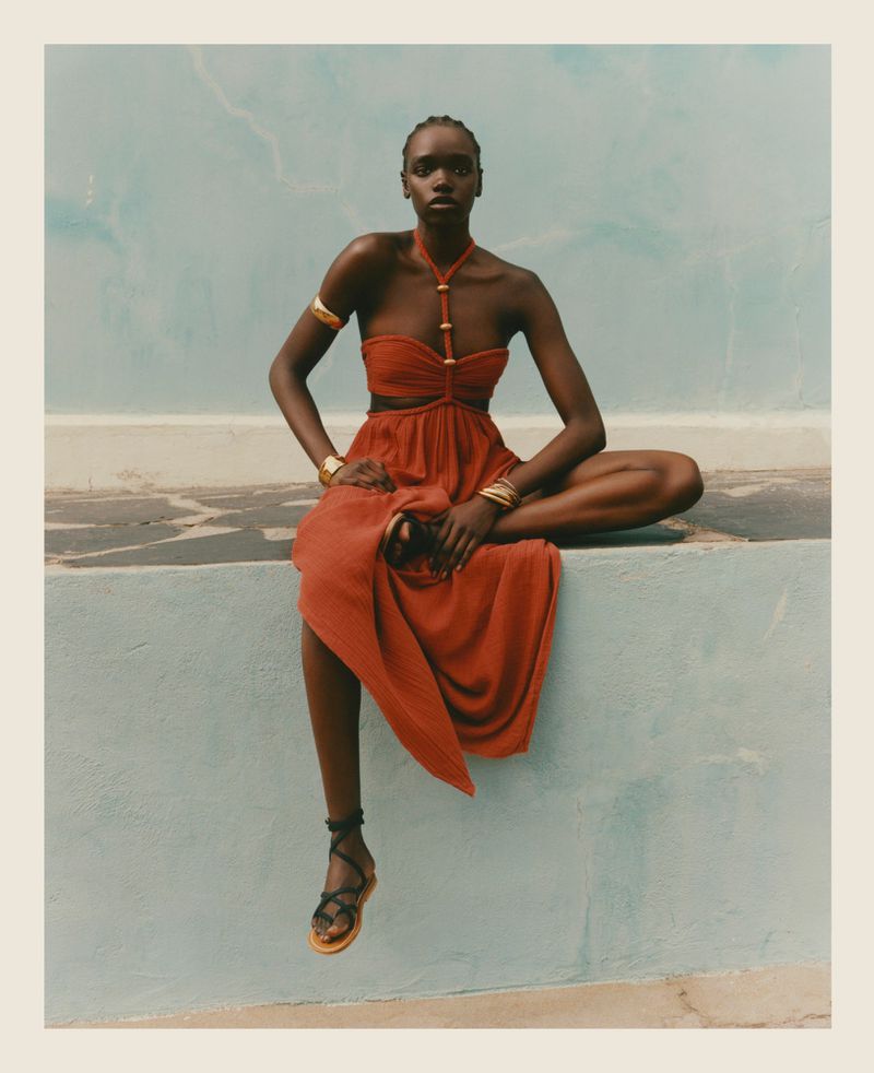 A single female model sits cross-legged with hands resting on her thighs, wearing a halter orange dress and gold bracelets against a blue wall.