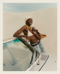 Smiling Black model in white bikini top and headscarf leaning against a classic car for Net-A-Porter SS25