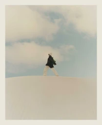 Model standing on a pale sand dune wearing a dark jacket, light trousers, and a beanie against an overcast sky.