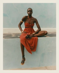 Model in a rust orange cut-out dress with gold jewelry seated against a pale blue wall for Net-A-Porter SS'25