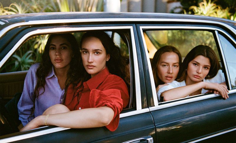 Four women are inside a dark-colored car, looking out of the windows. Two are in the front seat and two are in the back.