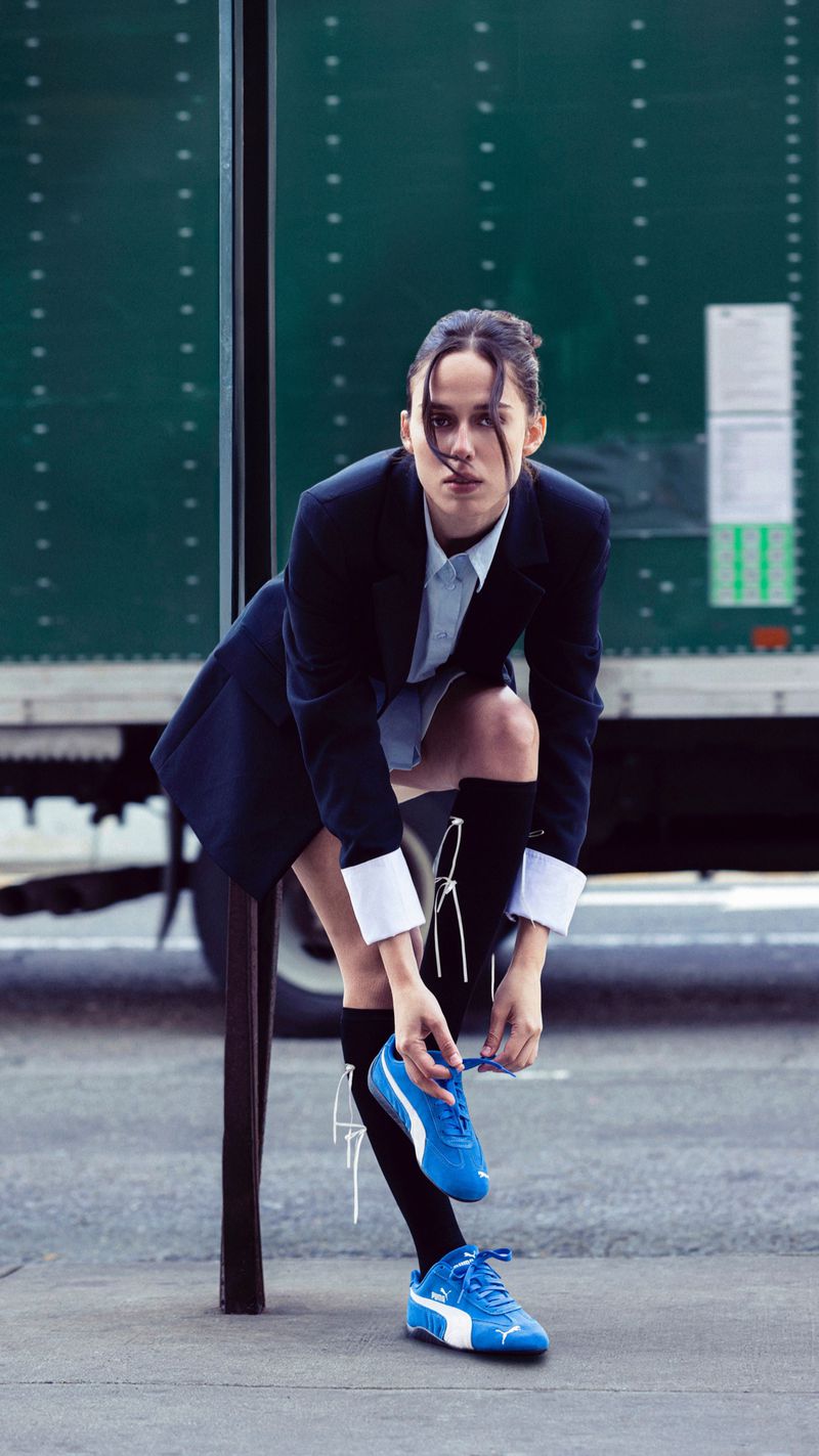 A stylish woman in a blazer and skirt ties her blue Puma sneakers in an urban setting with a green truck in the background.