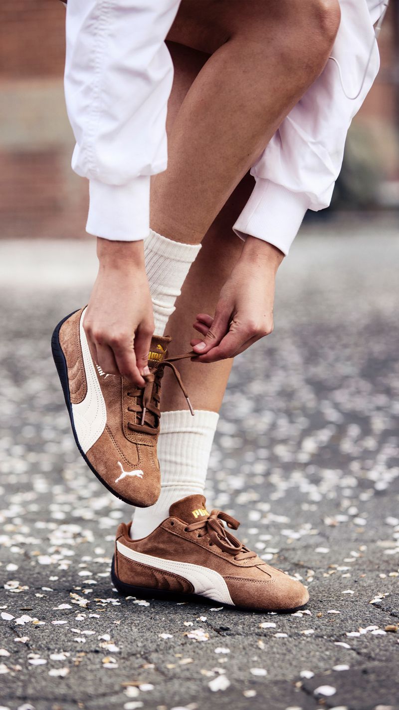 Person ties the shoelaces of brown and white Puma sneakers outdoors on pavement with scattered petals. Mid-level view, natural light.