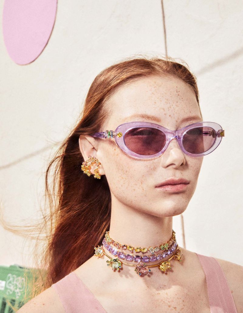 Close-up of a redhead woman wearing lilac sunglasses, jewelry, and a pink top, against a bright background, for a fashion concept.