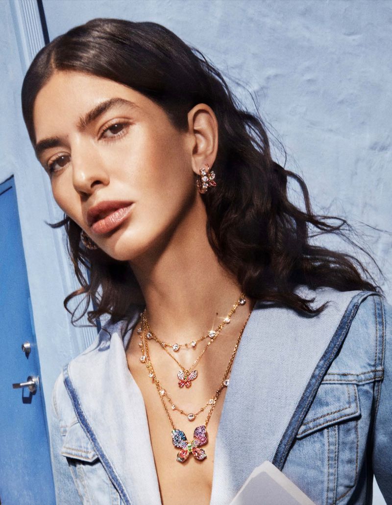 Brunette woman poses in a denim jacket with butterfly-themed layered necklaces and earrings against a light blue background.