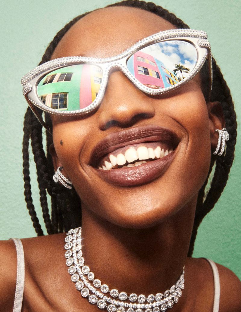 Close-up of a Black woman smiling, wearing sunglasses reflecting a colorful building and palm tree, along with jewelry against a green backdrop.