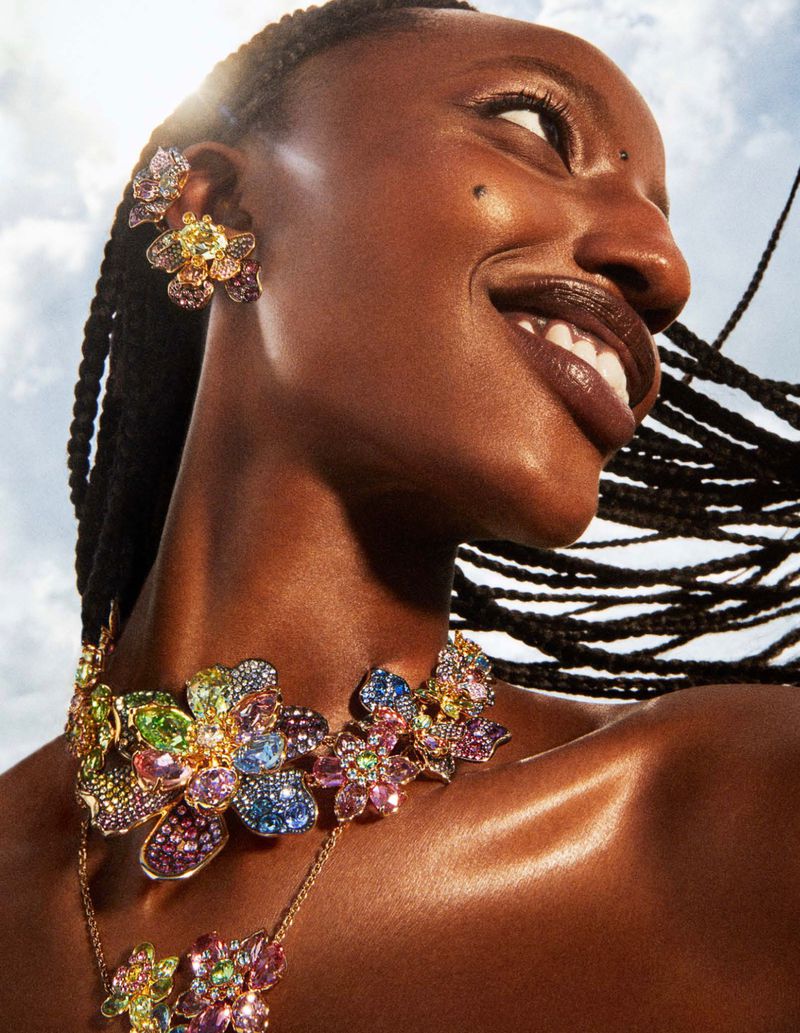 Close-up of a smiling Black woman showcasing jewelry, including a multi-colored flower necklace and earrings, against a blue sky backdrop.