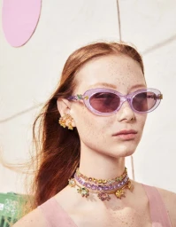 Close-up portrait of a woman wearing purple sunglasses, layered crystal necklaces, and crystal ear cuffs for Swarovski.
