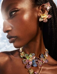 Close-up portrait of a model wearing vibrant floral crystal ear cuffs and necklace against a bright sky background.