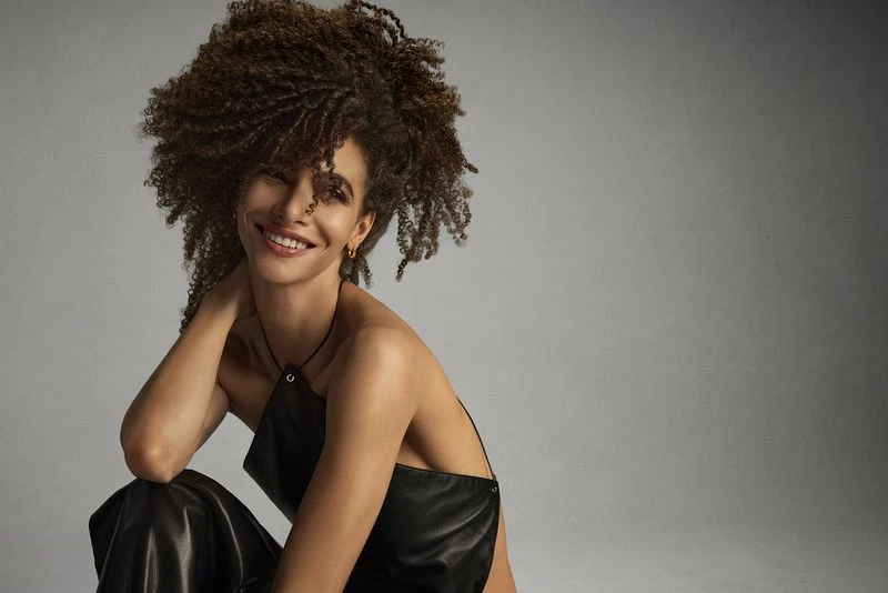 Portrait of a smiling woman with curly brown hair, wearing a black leather top and pants, posing in a photo studio against a gray backdrop.