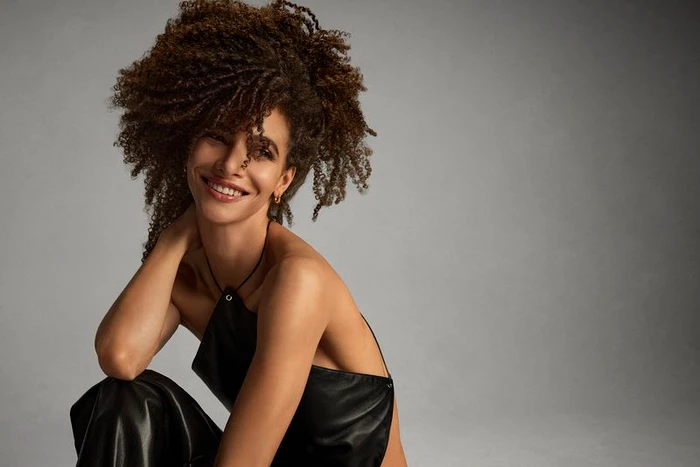 Woman with voluminous curly dark hair smiling, wearing a black top against a grey studio backdrop.