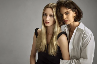 Two female models posing together, one with long blonde hair in a black top and one with dark bobbed hair in a white shirt.