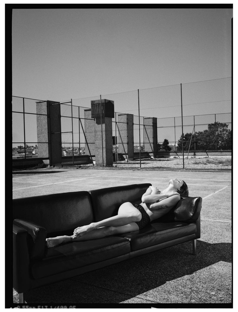 Black and white photo of a fair-skinned adult woman in underwear lounging on a dark couch on a tennis court, under a sunny sky.