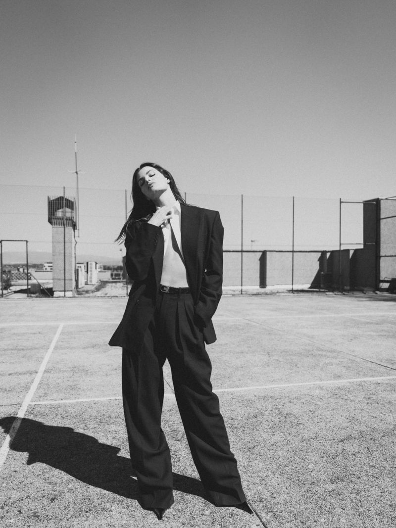 Black and white portrait of a female fashion model wearing a dark suit, posing on a rooftop, with an urban skyline and clear sky.