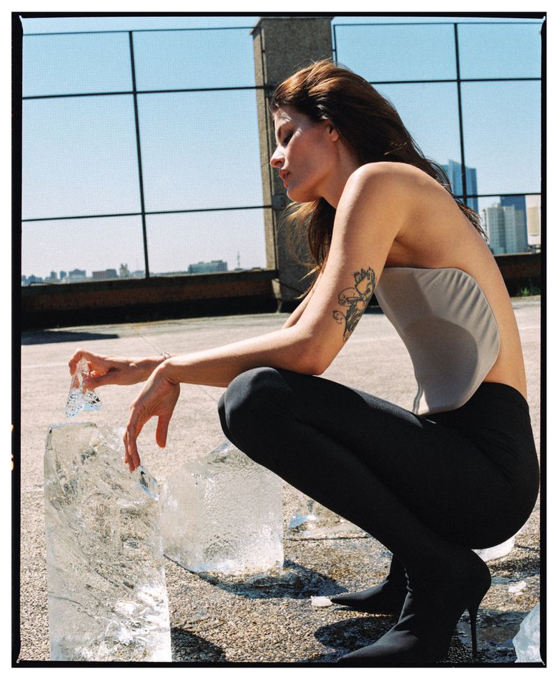 Woman squats next to ice blocks on an urban rooftop with city skyline background, wearing top, leggings, and stiletto sock boots.