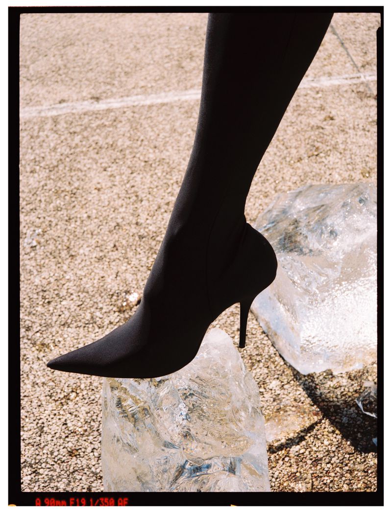 A black pointed-toe high-heel boot on a woman's leg rests on a block of ice against a neutral background. Fashion photography.
