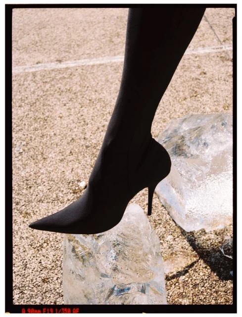 A black pointed-toe high-heel boot on a woman's leg rests on a block of ice against a neutral background. Fashion photography.