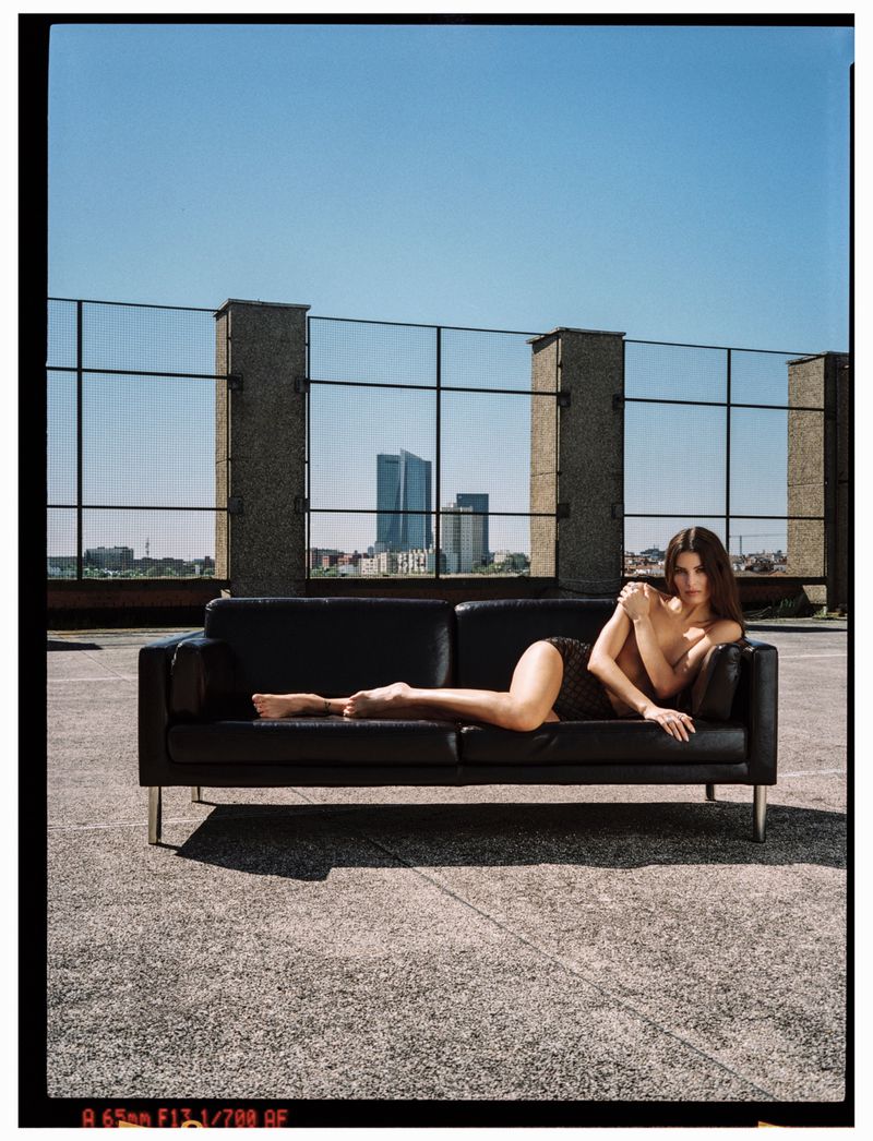 Adult female lying on a black couch on a rooftop with a city skyline background and clear blue sky in daytime, full body shot.