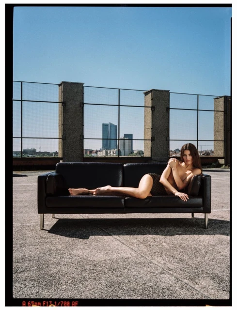 Adult female lying on a black couch on a rooftop with a city skyline background and clear blue sky in daytime, full body shot.