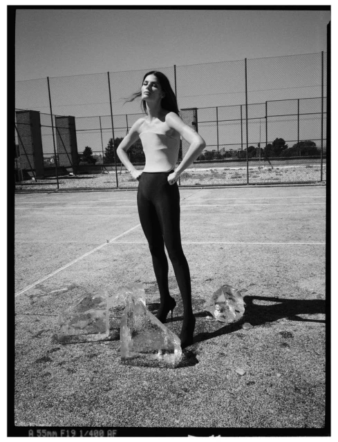 Monochrome fashion shot of a slender adult woman standing on an outdoor court with ice blocks at her feet under a bright sky.