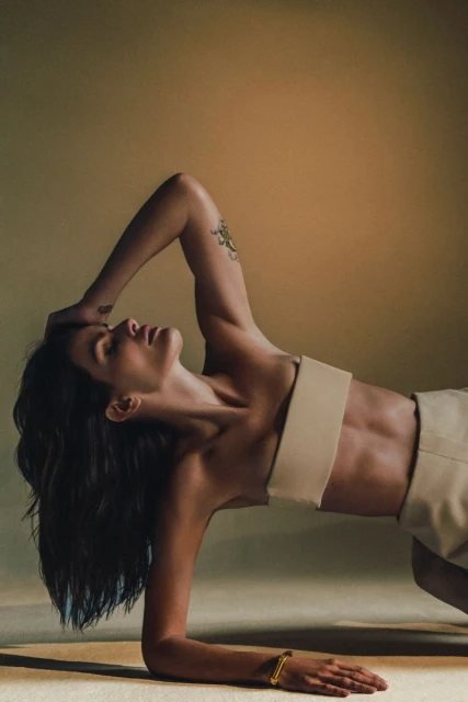 Portrait of an adult woman in a studio setting with beige background, wearing matching bra top and shorts. She's posed on the floor.