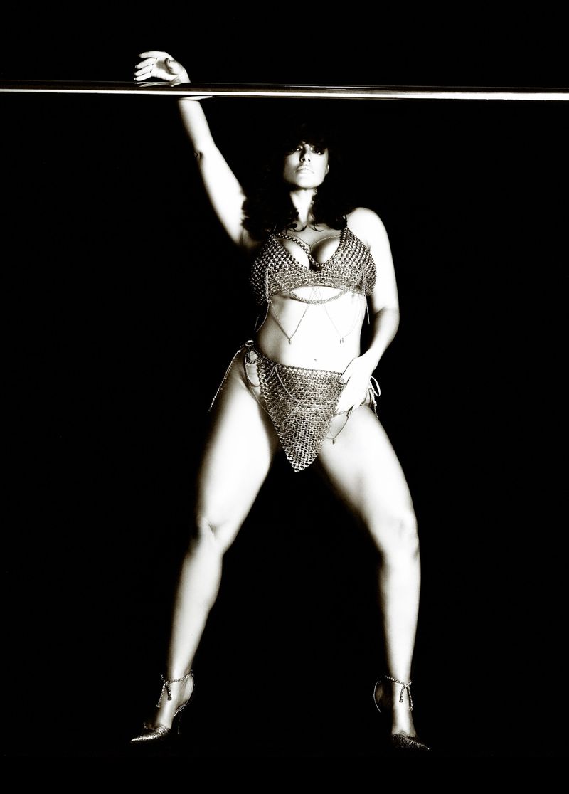 Monochrome image of a woman in chainmail-style lingerie, striking a pose against a dark background while grasping a metal bar overhead.