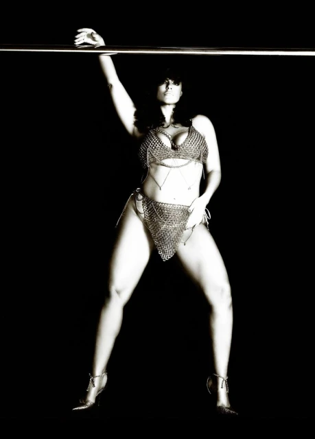 Monochrome image of a woman in chainmail-style lingerie, striking a pose against a dark background while grasping a metal bar overhead.