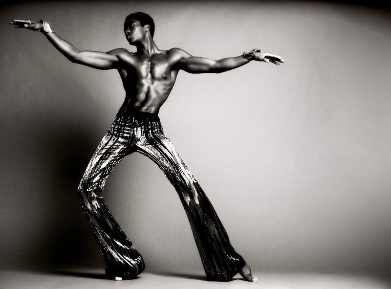 Black and white shot of a muscular African American man in patterned pants, posing with arms outstretched against a plain background.