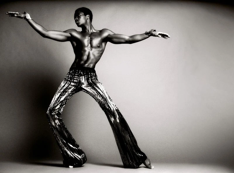 Black and white shot of a muscular African American man in patterned pants, posing with arms outstretched against a plain background.