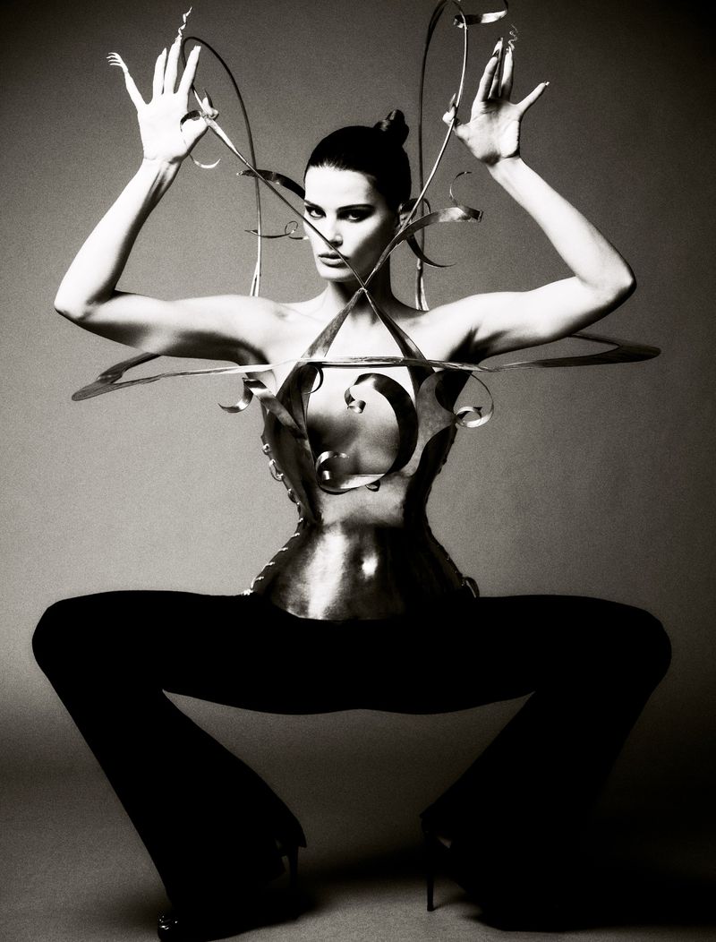 Black and white fashion shot of an adult female model wearing a metallic corset and posing with arms raised against a neutral backdrop.