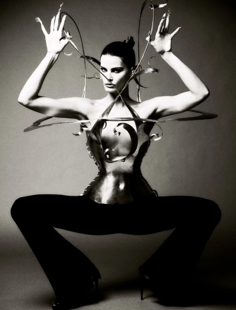Black and white fashion shot of an adult female model wearing a metallic corset and posing with arms raised against a neutral backdrop.