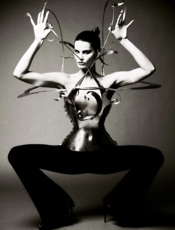 Black and white fashion shot of an adult female model wearing a metallic corset and posing with arms raised against a neutral backdrop.