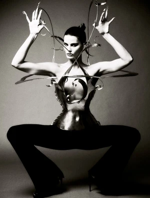 Model wearing metallic sculptural bodice and dark trousers posing with hands raised against a grey seamless background
