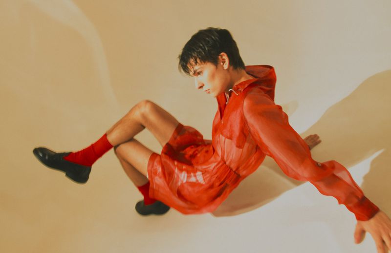 Fashion shot of a young adult male model wearing a red hooded outfit with black shoes, posing on a beige background with shadow.