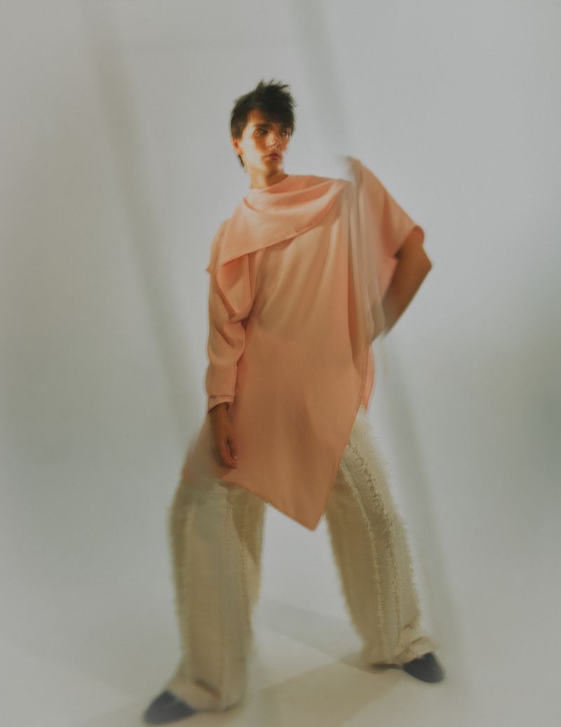A stylish person poses indoors wearing a salmon pink tunic and wide-legged pants, against a white backdrop with soft, diffused lighting.