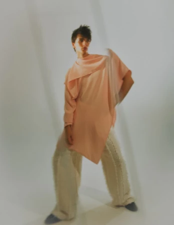 A stylish person poses indoors wearing a salmon pink tunic and wide-legged pants, against a white backdrop with soft, diffused lighting.