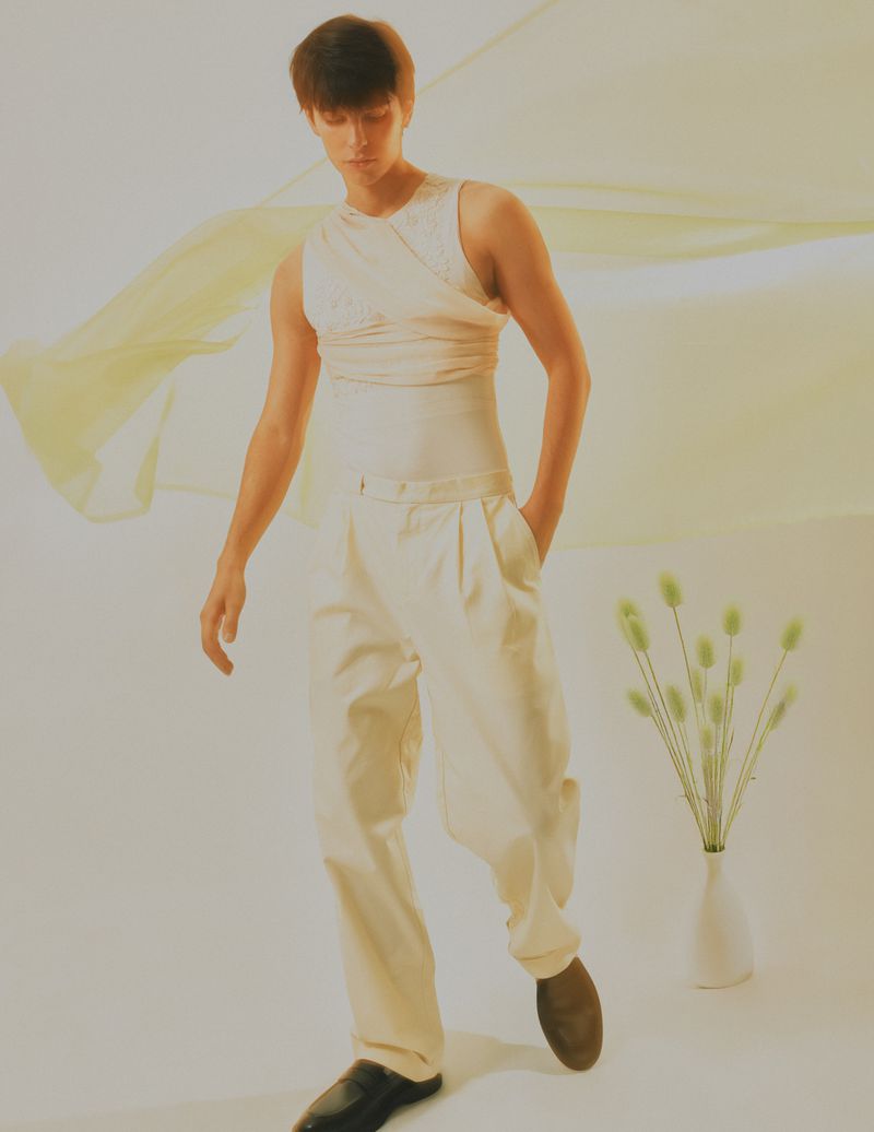 Studio fashion portrait of a young adult male in neutral tones, wearing a sleeveless top and slacks, posed in front of a neutral backdrop.