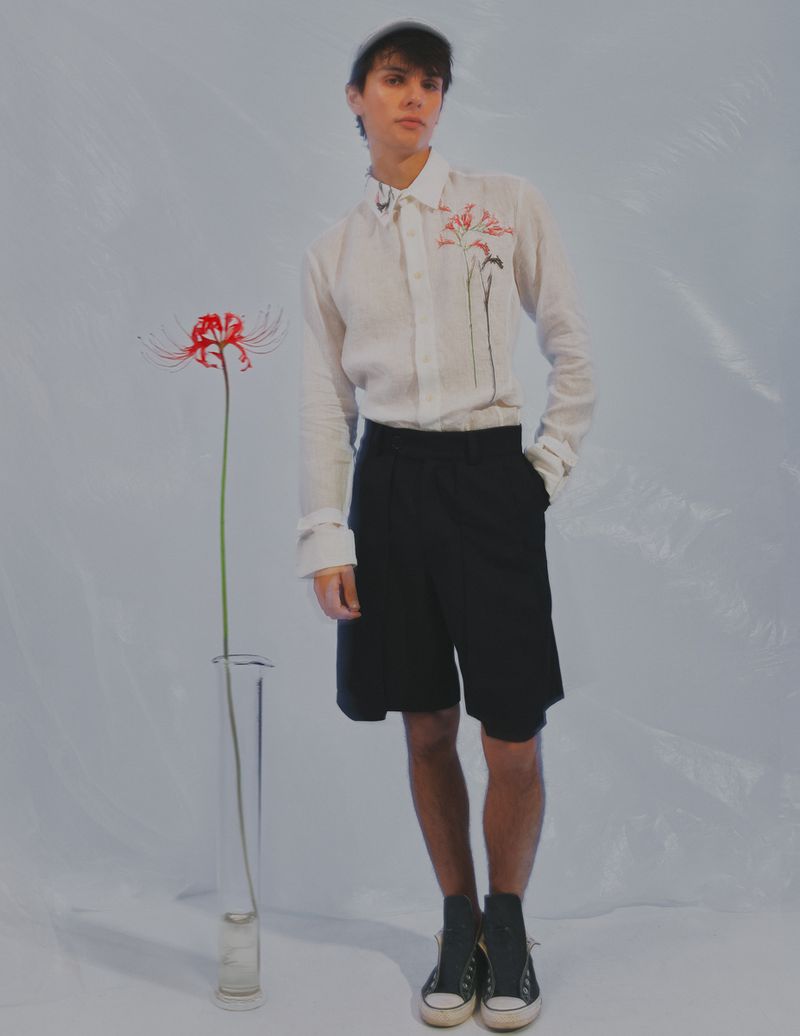 Young man in white shirt and black shorts poses in front of a spider lily in a clear vase, minimalist, fashion-forward.