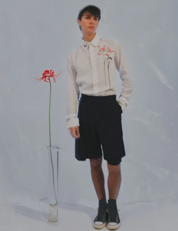 Young man in white shirt and black shorts poses in front of a spider lily in a clear vase, minimalist, fashion-forward.