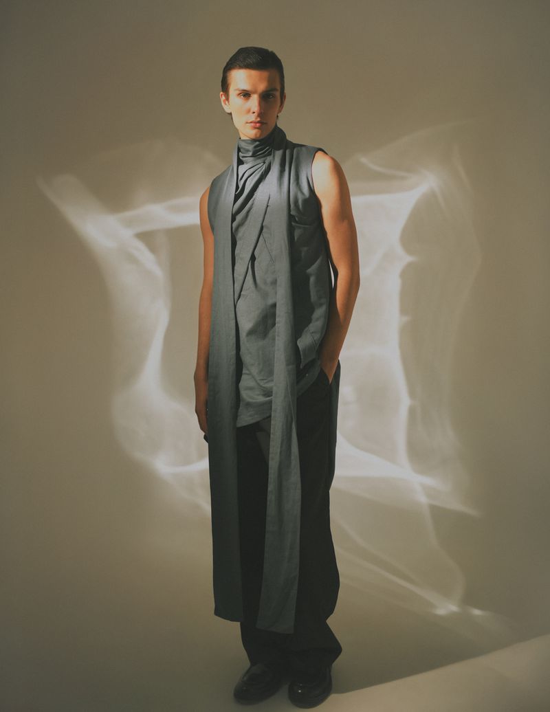 Studio portrait of a young adult male in fashionable gray sleeveless top and black pants posing against soft patterned background.