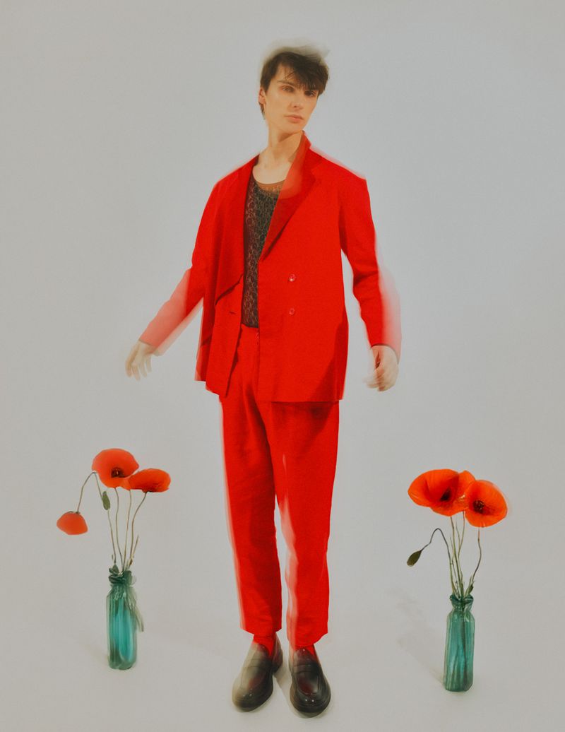 A slender person in a bright red suit stands in a studio setting with two vases of red poppies, a clean and striking fashion image.