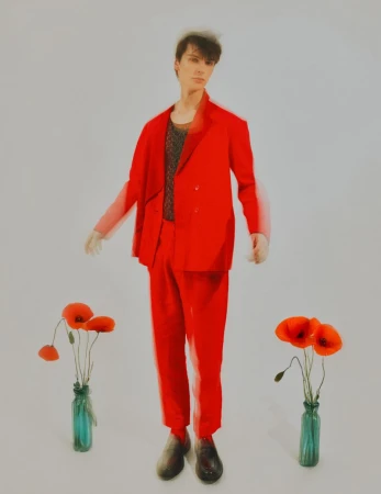 A slender person in a bright red suit stands in a studio setting with two vases of red poppies, a clean and striking fashion image.
