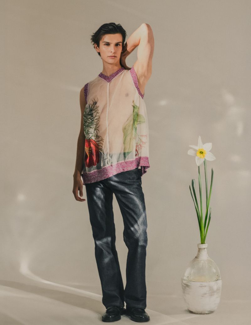 A fashionable male model poses in a semi-sheer top and faux-leather pants next to a daffodil in a glass vase in a studio setting.