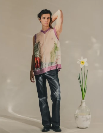 A fashionable male model poses in a semi-sheer top and faux-leather pants next to a daffodil in a glass vase in a studio setting.
