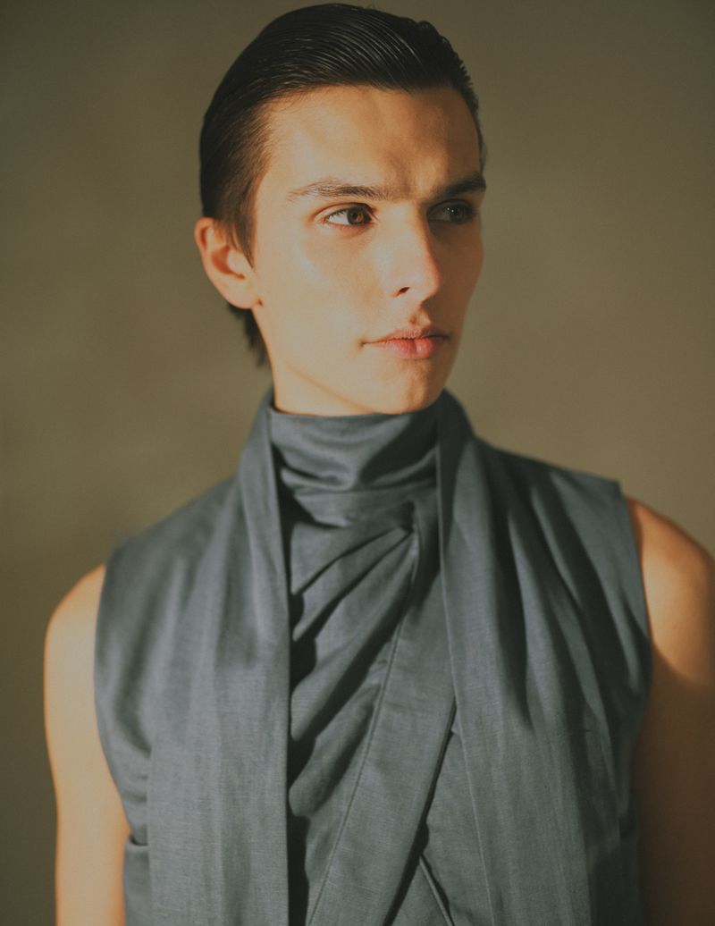 Portrait of an androgynous adult male in a gray sleeveless tunic, softly lit against a neutral background in a studio setting.