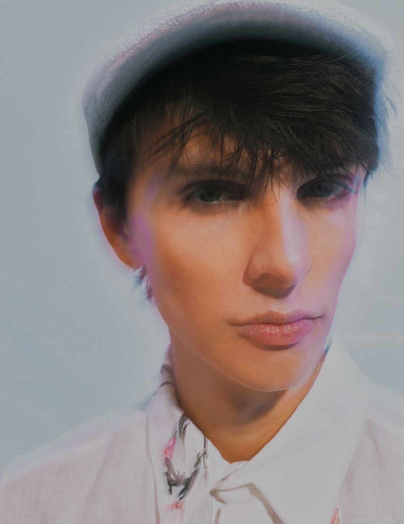Close-up of a person with dark hair wearing a cap and white shirt with abstract print against a soft, blurred background.