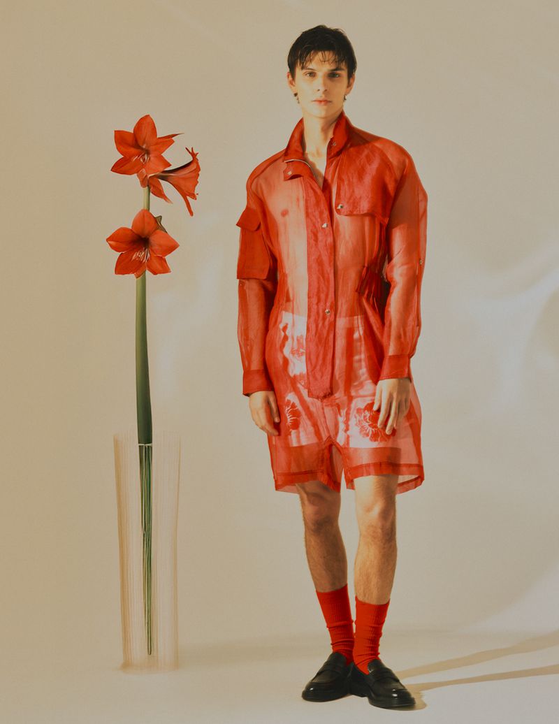 Fashion shot of a young adult man wearing a semi-sheer red ensemble next to a vase of red amaryllis flowers on a neutral background.