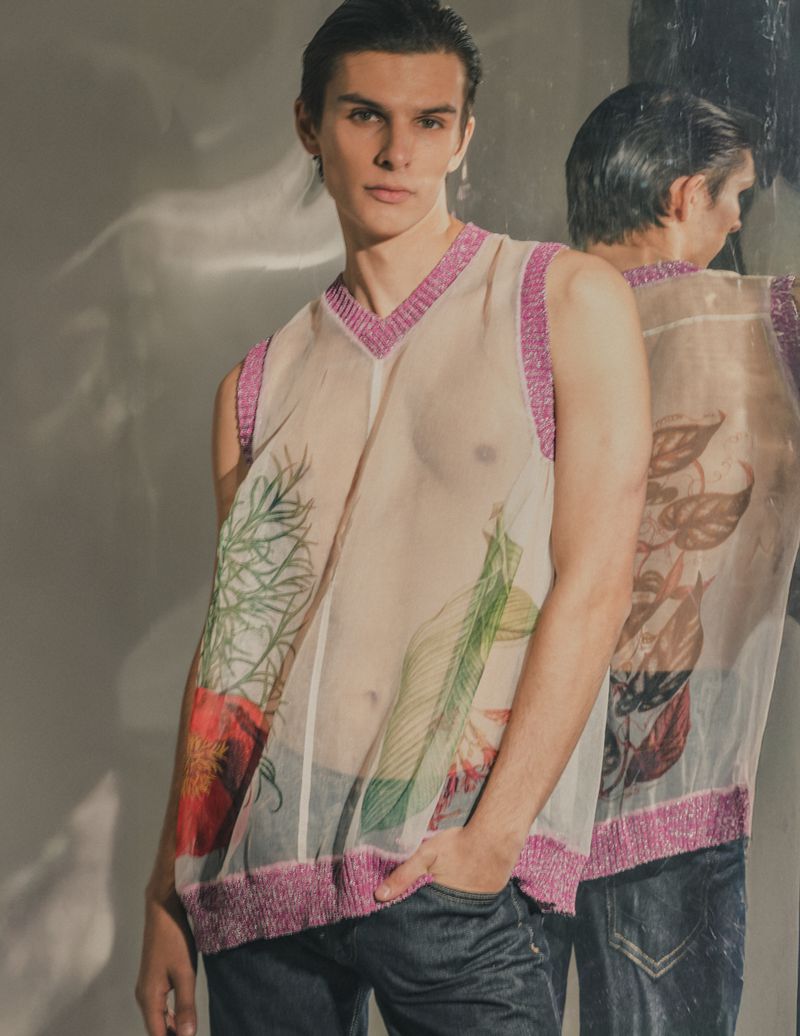 A young male model poses in a sheer floral print top and jeans with his reflection behind him in a fashion photography studio setting.